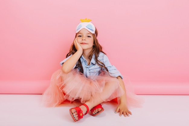 Bambina allegra in gonna di tulle seduto sul pavimento bianco isolato su sfondo rosa. Bella principessa bambino con maschera sulla testa sorridendo alla telecamera, esprimendo la felicità del bel ragazzo