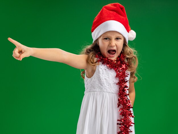 Bambina arrabbiata che indossa il cappello di Natale con la ghirlanda sui punti del collo sul lato isolato su sfondo verde