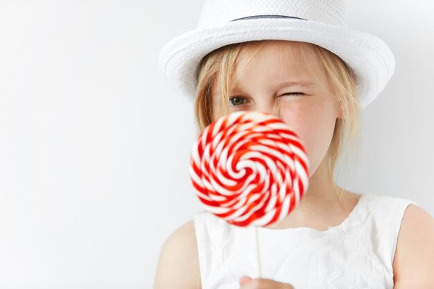 Bambina bionda che indossa un cappello bianco e tenendo la caramella di zucchero