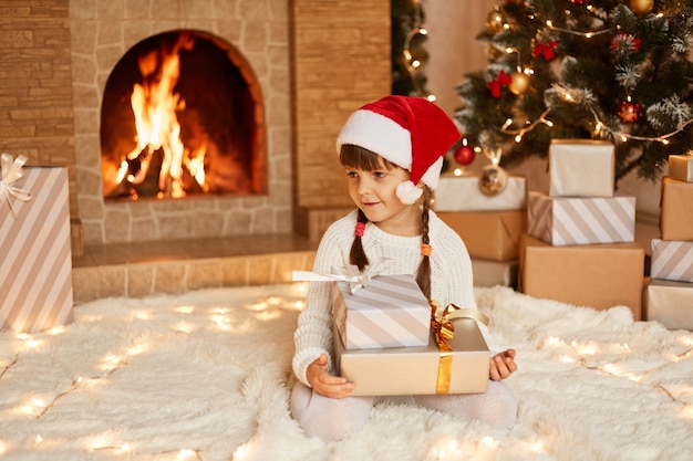 Bambina carina positiva che indossa maglione bianco e cappello di babbo natale, con in mano una pila di regali, seduta sul pavimento su un tappeto morbido vicino all'albero di Natale, scatole regalo e camino.