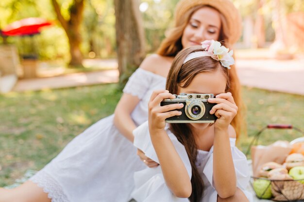 Bambina castana con il nastro nei capelli che cattura foto della natura che gode del fine settimana. Outdoor ritratto di affascinante giovane donna nel parco con la figlia che tiene la macchina fotografica.