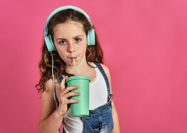 Bambina che ascolta musica con le cuffie e beve un succo su uno sfondo rosa