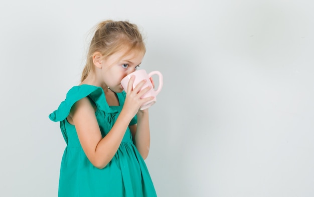 Bambina che beve tazza di tè in vestito verde e che sembra assetata. .