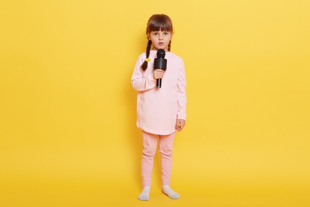 Bambina che canta una canzone con cam e espressione facciale seria, guarda la telecamera con sguardo preoccupato, essendo confusa per organizzare la performance, indossando abbigliamento casual, isolato su sfondo giallo.