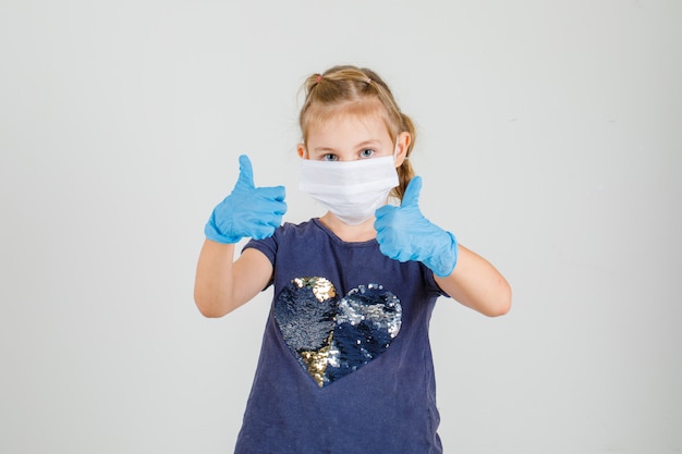 Bambina che compone i pollici in maglietta, guanti e maschera medica e che sembra sicura. vista frontale.