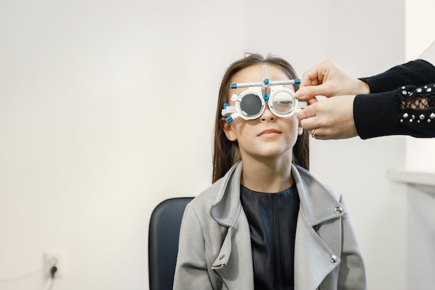 Bambina che controlla la sua vista al centro di oftalmologia. Ottico optometrista femminile che esegue test della vista. Ragazza bruna che indossa giacca grigia e camicia nera.