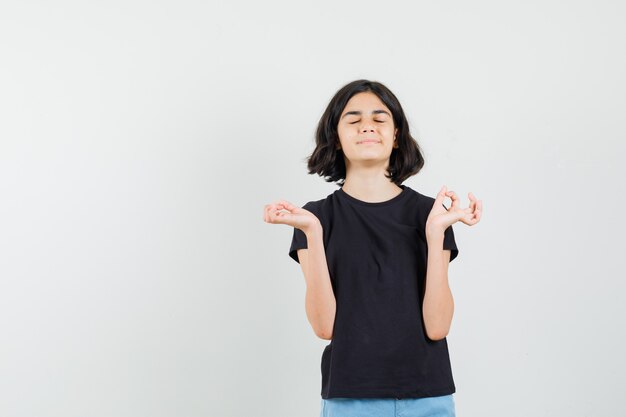 Bambina che fa meditazione con gli occhi chiusi in maglietta nera, pantaloncini e cerca pacifica, vista frontale.