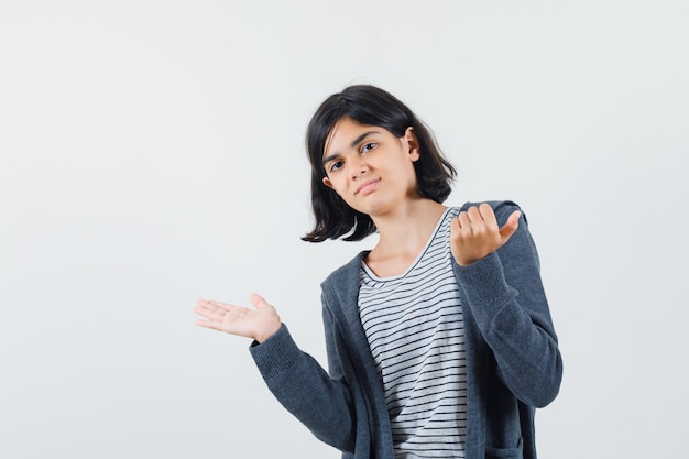 Bambina che finge di mostrare qualcosa in t-shirt, giacca e sembra sicura.