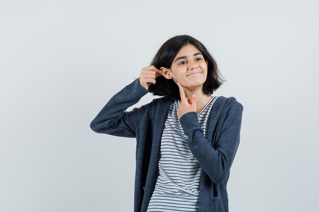 Bambina che indica al suo orecchio in t-shirt, giacca e sembra allegra,