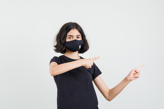 Bambina che indica di lato in maglietta nera, maschera e guardando fiducioso, vista frontale.