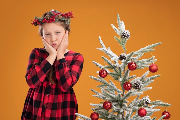 Bambina che indossa una ghirlanda di natale in camicia a quadri felice e positiva in piedi accanto a un albero di natale sul muro arancione orange