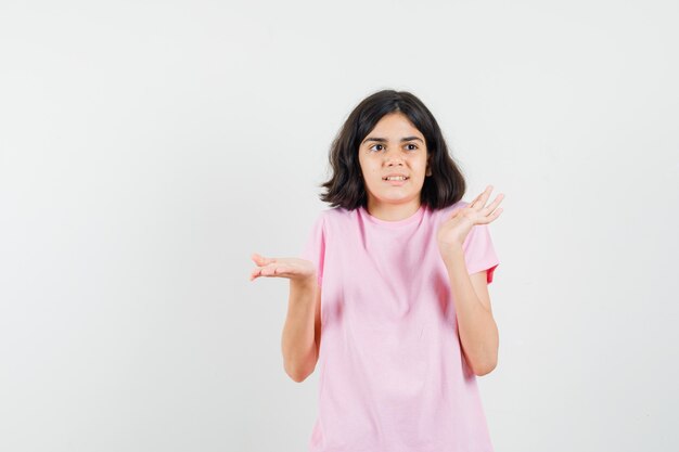 Bambina che mostra gesto impotente alzando le spalle in maglietta rosa e guardando confuso, vista frontale.