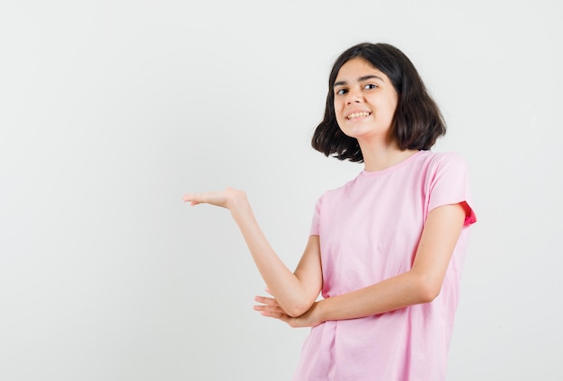 Bambina che mostra qualcosa o dà il benvenuto in maglietta rosa e sembra allegra, vista frontale.
