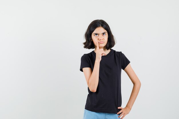 Bambina che osserva in su in maglietta nera, pantaloncini e guardando pensieroso, vista frontale.