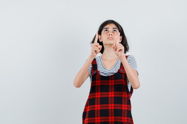 Bambina che osserva in su mentre indica verso l'alto in t-shirt, tuta e guardando paziente,