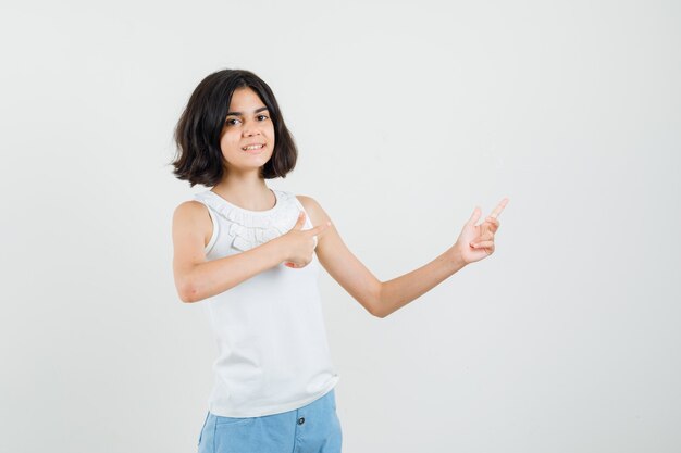 Bambina che punta di lato e sorridente in camicetta bianca, pantaloncini, vista frontale.