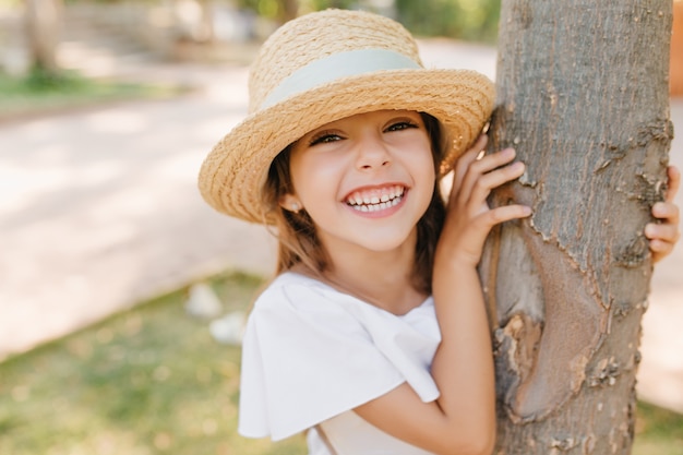 Bambina che ride con la pelle leggermente abbronzata in posa nel parco toccando l'albero. Ritratto di close-up all'aperto di allegro ragazzo dai capelli scuri in cappello vintage con nastro divertendosi in giardino.
