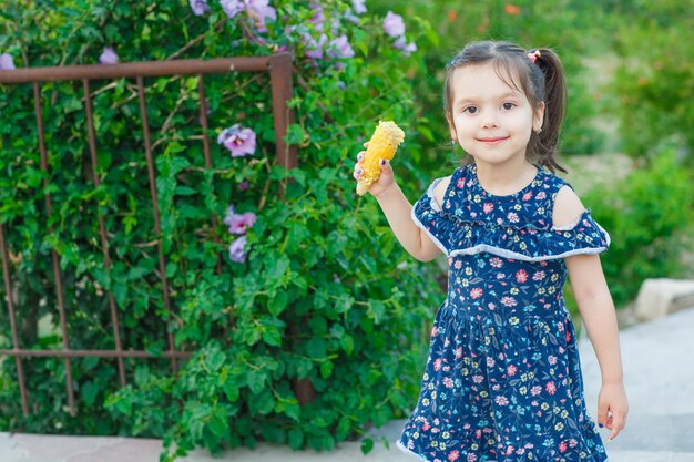 Bambina che tiene il mais e che guarda l'obbiettivo in giardino in abito primaverile e guardando felice. vista frontale.