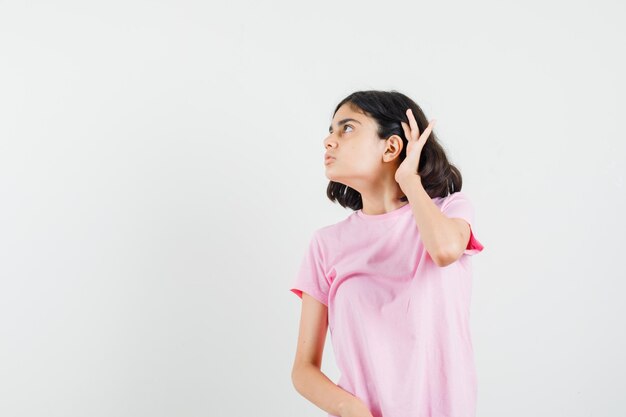 Bambina che tiene la mano dietro l'orecchio in maglietta rosa e guardando curioso, vista frontale.