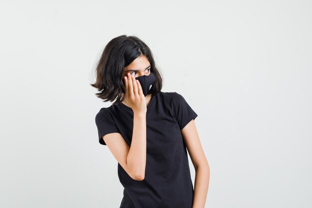 Bambina che tiene la mano sulla guancia in maglietta nera, maschera e sguardo curioso, vista frontale.
