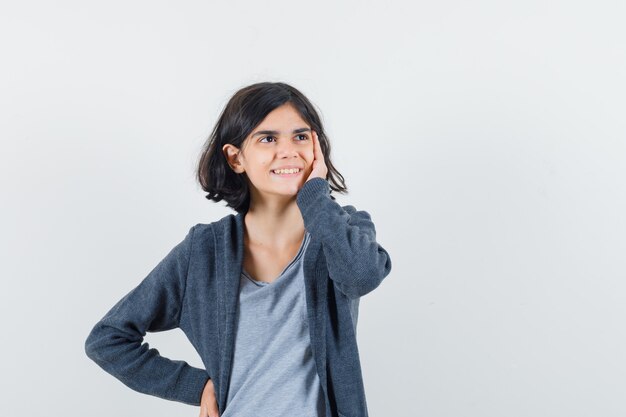 Bambina che tiene la mano sulla guancia in t-shirt, giacca e dall'aspetto sognante, vista frontale.