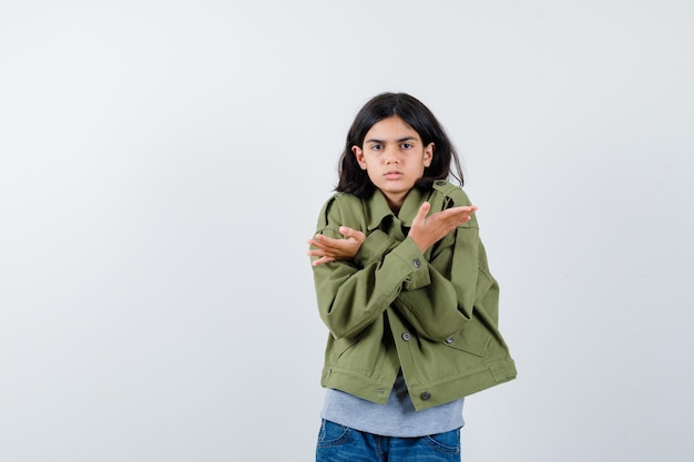 Bambina che tiene le braccia incrociate sul petto in cappotto, t-shirt, jeans e sembra esitante, vista frontale.