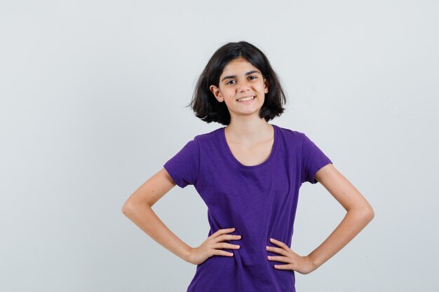 Bambina che tiene le mani sulla vita in maglietta e sembra gioiosa,