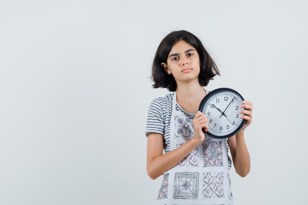 Bambina che tiene orologio da parete in t-shirt, grembiule e che sembra calma,