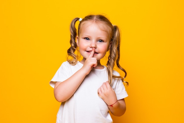 Bambina con il dito sulla bocca che dice Shh isolato sopra la parete gialla