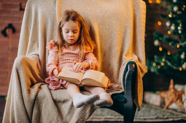 Bambina con il libro che si siede nella sedia dall&#39;albero di Natale