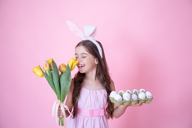 Bambina con le orecchie del coniglietto di Pasqua e un vassoio di uova nelle sue mani annusando un mazzo di tulipani su una parete rosa.