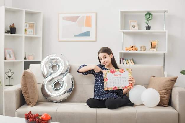 Bambina contenta per la felice festa della donna che tiene e indica il calendario seduta sul divano nel soggiorno