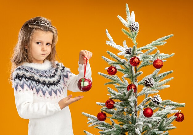 Bambina di pensiero che sta vicino all'albero di Natale che indossa il diadema con la ghirlanda sulla tenuta del collo e punti alla palla di Natale isolata su fondo arancio