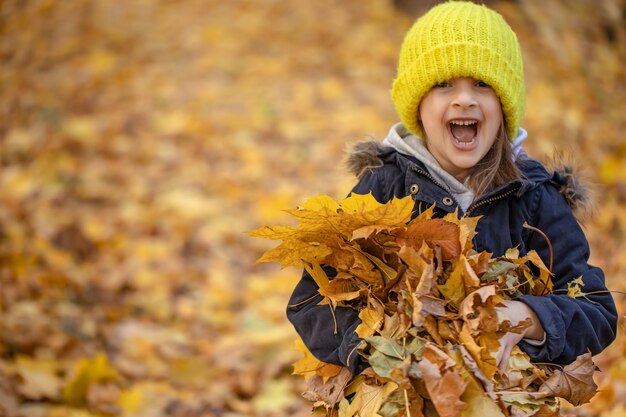 Bambina divertente che tiene un mucchio di foglie autunnali nella foresta su uno sfondo sfocato, copia dello spazio.