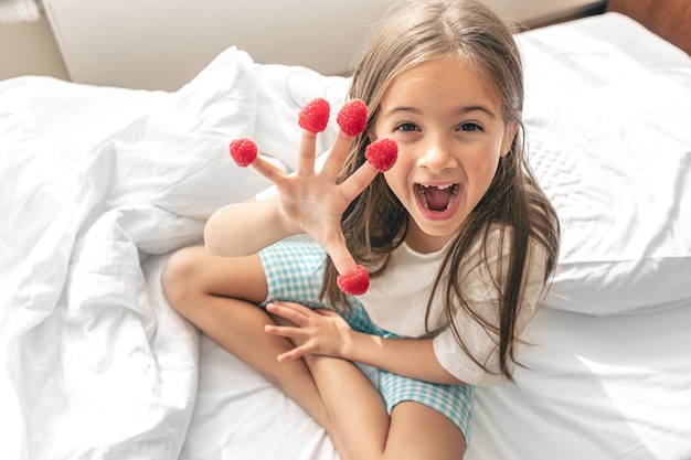 Bambina divertente con lamponi sulle dita a letto al mattino