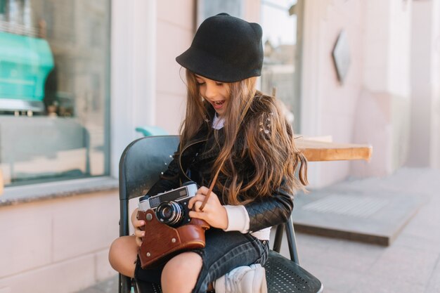 Bambina eccitata dai capelli lunghi che si siede sulla sedia nel caffè all'aperto e che gioca con la retro macchina fotografica. Ritratto all'aperto del bambino riccio interessato che aspetta il fotografo della madre che tiene la sua roba.