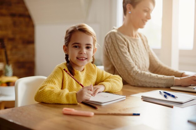Bambina felice che disegna mentre sua madre sta lavorando al computer portatile a casa