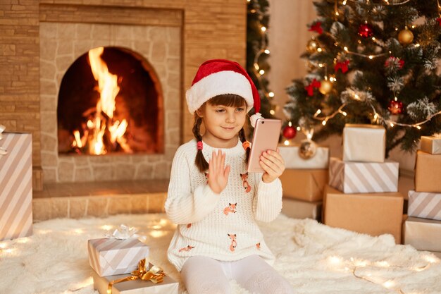 Bambina felice che indossa un maglione bianco e cappello di babbo natale, in posa in una stanza festiva con camino e albero di Natale, agitando la mano alla fotocamera del cellulare, con una videochiamata.