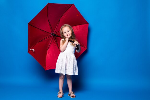 Bambina felice con l'ombrello rosso che posa sulla parete blu.