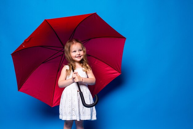 Bambina felice con l'ombrello rosso che posa sulla parete blu.