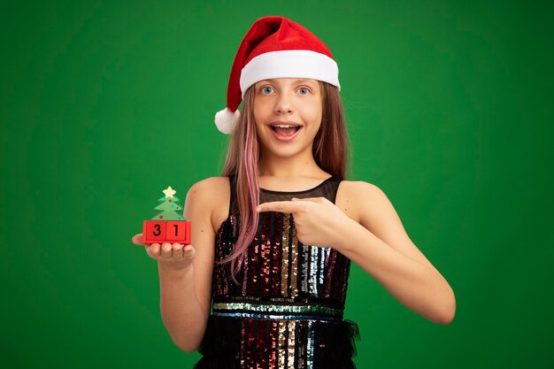 Bambina felice e sorpresa in abito da festa glitter e cappello da Babbo Natale che mostra cubi giocattolo con la data di capodanno che punta con il dito indice sorridendo allegramente in piedi su sfondo verde