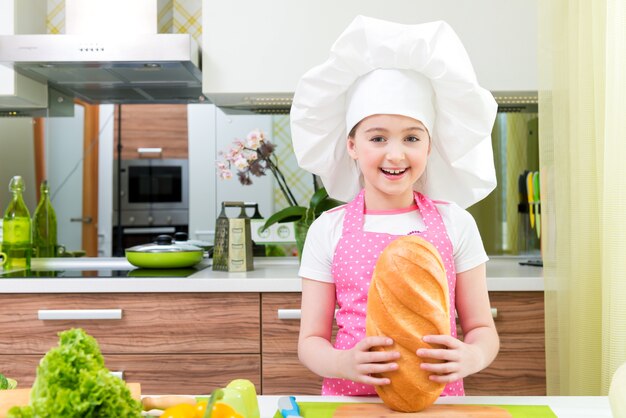 Bambina felice in grembiule rosa con pane nelle sue mani in cucina.