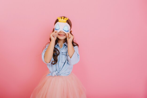 Bambina graziosa con capelli lunghi del brunette in gonna di tulle che tiene la maschera di sonno con gli occhi azzurri e corona d'oro isolato su sfondo rosa. Celebrare la festa di compleanno, divertirsi al carnevale per i bambini