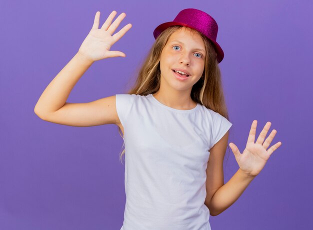 Bambina graziosa in cappello di festa che alza le palme sorridenti felici e positive, concetto della festa di compleanno che sta sopra fondo viola