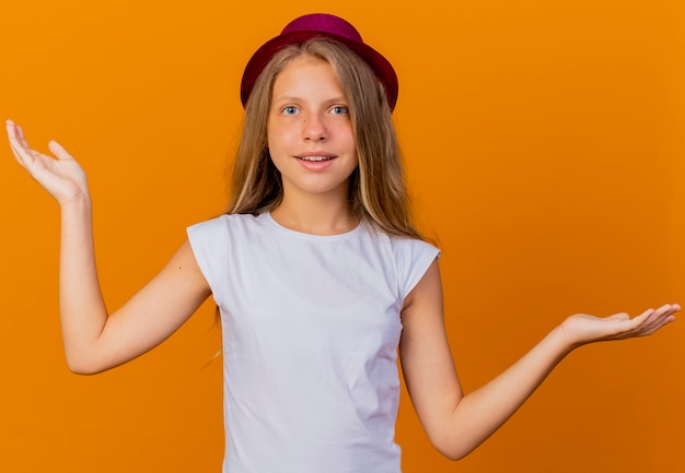 Bambina graziosa in cappello di festa che guarda l'obbiettivo sorridente diffondendo le braccia ai lati, concetto di festa di compleanno in piedi su sfondo arancione
