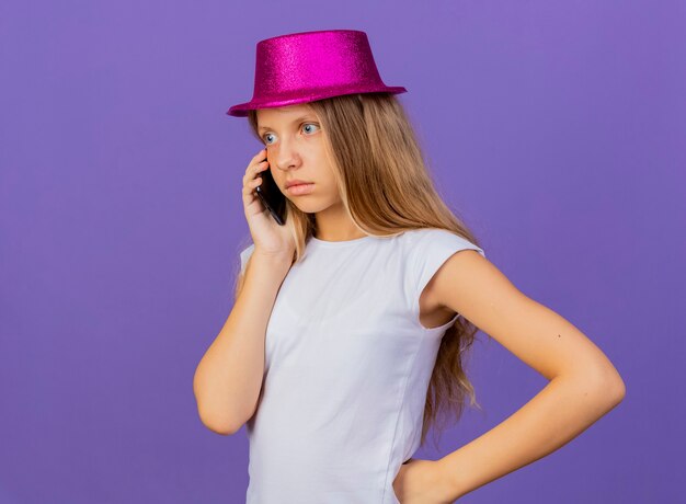 Bambina graziosa in cappello di festa che parla sul telefono cellulare che è confuso, concetto di festa di compleanno in piedi su sfondo viola