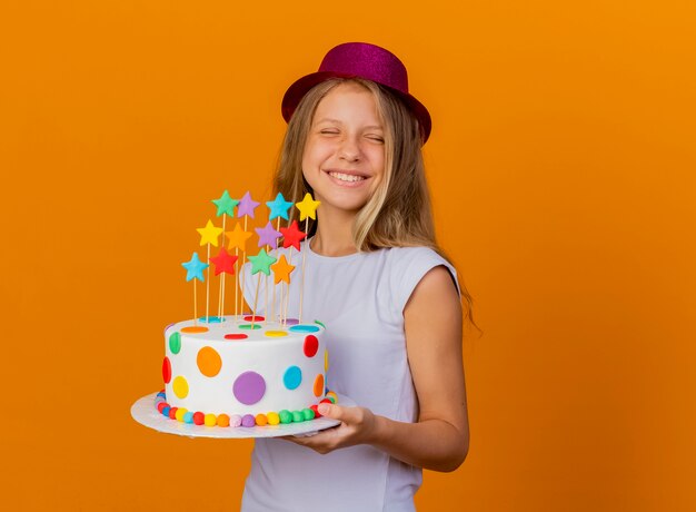 Bambina graziosa in cappello di festa che tiene cakehapy di compleanno ed eccitato, concetto della festa di compleanno che sta sopra fondo arancio