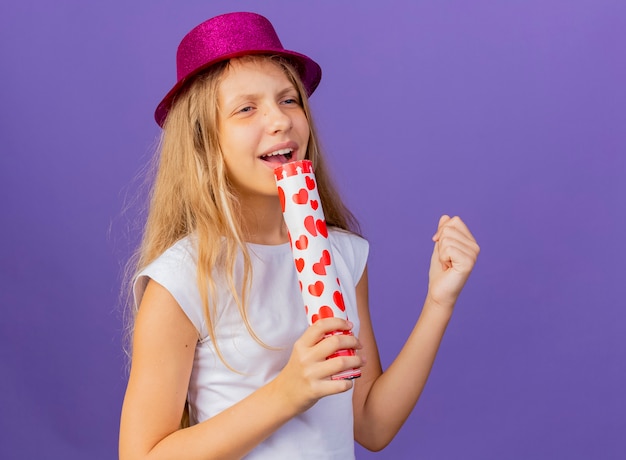 Bambina graziosa in cappello di festa che tiene il cracker del partito usando come canto del microfono, concetto della festa di compleanno che sta sopra fondo viola