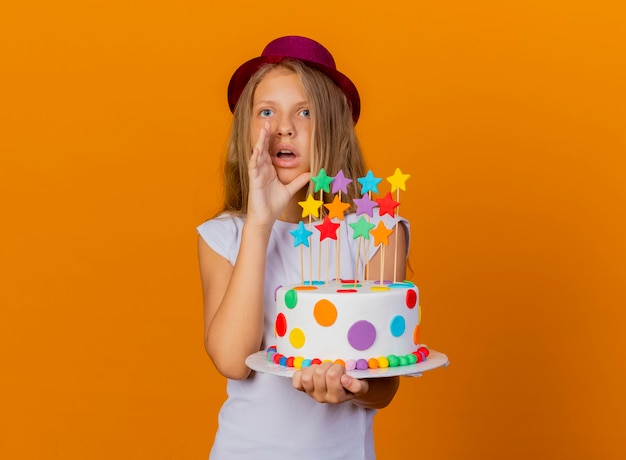 Bambina graziosa in cappello di festa che tiene la torta di compleanno che sembra sorpresa che chiama qualcuno, concetto della festa di compleanno