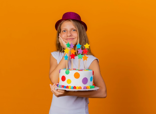 Bambina graziosa in cappello di festa che tiene la torta di compleanno che sorride con il fronte felice, concetto della festa di compleanno